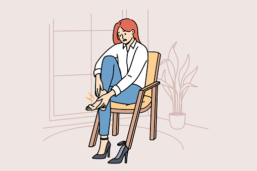Woman with varicose veins feels pain in legs due to uncomfortable high-heeled shoes, sits on chair in apartment. Girl needs medicinal ointment to cure varicose veins or calluses on heel