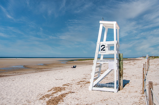 A white wooden lifeguard chair overlooks the wide expanse of sand at low tide where only two people relax at Chapin Beach  in Dennis, Massachusetts on a May afternoon.