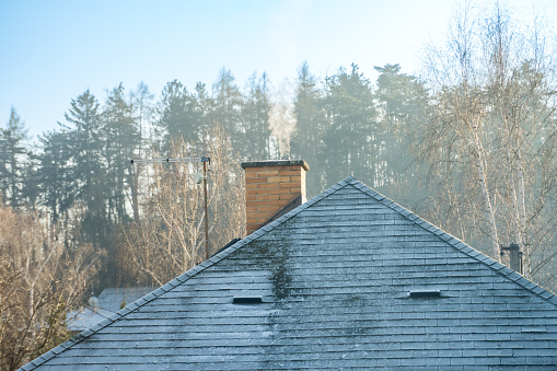 Smoke rises from the chimney on the house. Roof with smoking chimney and trees in winter. High quality photo