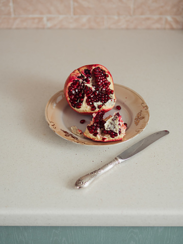 An open pomegranate rests on a plate against a white table, providing a striking contrast against the soft pastel pink wall. The intricate details of the pomegranate's vibrant red seeds are accentuated in the foreground. Adding a touch of color diversity, a portion of a turquoise-colored drawer is visible in the frame, introducing a complementary hue to the elegant and harmonious composition of this kitchen setting. The combination of vivid red, neutral tones, and the splash of turquoise creates a visually appealing and balanced scene.