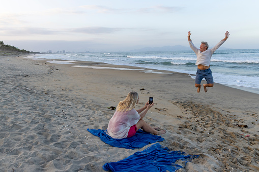 Mature woman takes photo of man jumping on beach at sunset
