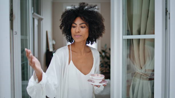 istock Curly woman touching rain at window. African lady looking camera holding coffee 1945732472