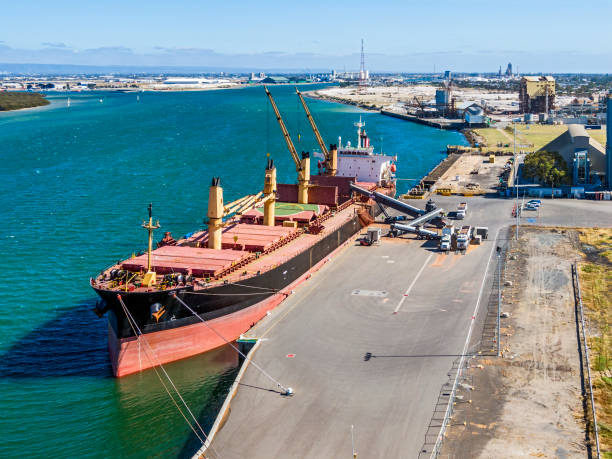 Aerial view, looking down on unrecognisable dock workers loading grain directly from trucks to ship via a series of mechanical conveyor belts in river port (Port Adelaide). ID, logos and flag all edited. Three semi-trucks are pictured at the loading point with grain spillage on the asphalt loading dock. Port infrastructure visible in background along with distant hill range.
