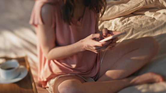 Closeup hands typing smartphone screen. Smiling girl resting in bed messaging online on cozy weekend. Relaxed woman using mobile phone enjoying hotel breakfast in pajamas. Carefree female in sunlight