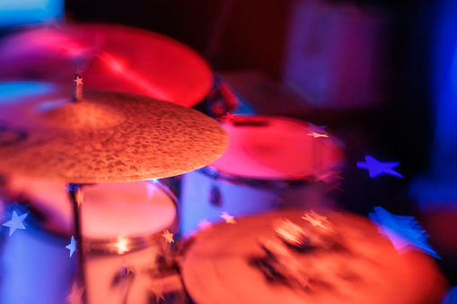 This captivating close-up image showcases a professional drum kit basked in the dramatic glow of red stage lighting, highlighting the detailed craftsmanship of the percussion instruments. The photograph captures the essence of a live music performance, with the chrome hardware of the drum rims and the textured surfaces of the drum heads gleaming under the vibrant lights. The ambient blue lighting in the background adds depth and contrast to the composition. Perfect for conveying the dynamic atmosphere of a concert or music venue, this image is ideal for articles, advertisements, and stories related to music performances, stage setups, and the passion behind the rhythm section of a band.