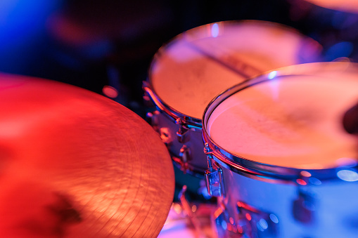 This captivating close-up image showcases a professional drum kit basked in the dramatic glow of red stage lighting, highlighting the detailed craftsmanship of the percussion instruments. The photograph captures the essence of a live music performance, with the chrome hardware of the drum rims and the textured surfaces of the drum heads gleaming under the vibrant lights. The ambient blue lighting in the background adds depth and contrast to the composition. Perfect for conveying the dynamic atmosphere of a concert or music venue, this image is ideal for articles, advertisements, and stories related to music performances, stage setups, and the passion behind the rhythm section of a band.