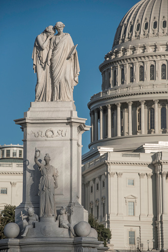 The Peace Monument, completed in 1878, stands 44 feet high in the northwestern quadrant of the circle to the west of the U.S. Capitol Building. It is dedicated to naval deaths during the Civil War.\n\nAt the top of the monument, a classically draped female figure representing Grief cries against the shoulder of History. History has inscribed on the tablet she holds, “They died that their country might live.” \n\nAnother classically-dressed female figure, Victory, stands below the other two, holding aloft a laurel wreath. At her feet are two small children, representing Mars and Neptune, the gods of war and the sea, respectively. \n\nThe monument is the work of sculptor Franklin Simmons (1839-1913).\n\nIn the background is the west face of the Capitol Building.