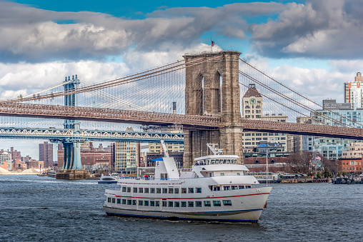 A tour boat passes beneath the Brooklyn Bridge on a day with blue sky and dramatic clouds. The Manhattan Bridge is visible in the background, as is DUMBO and adjacent sections of Brooklyn.