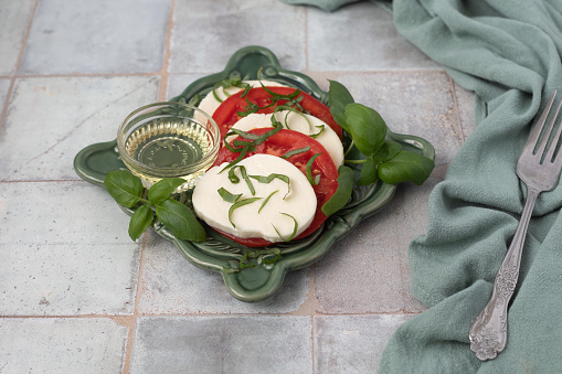 Fresh Caprese Salad with tomatoes, mozzarella cheese, and fresh basil on a green plate. Small bowl of olive oil.  Dark green background.