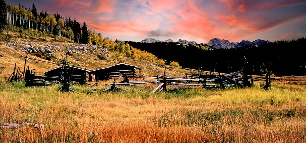 A deserted log cabin with a corral sits in a meadow making a picturesque scene. The colorful sunset sky adding to the scenic landscape.