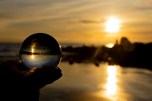 A close up image of a hand holding a photography glass lens ball against a golden sunset along the ocean shoreline.
