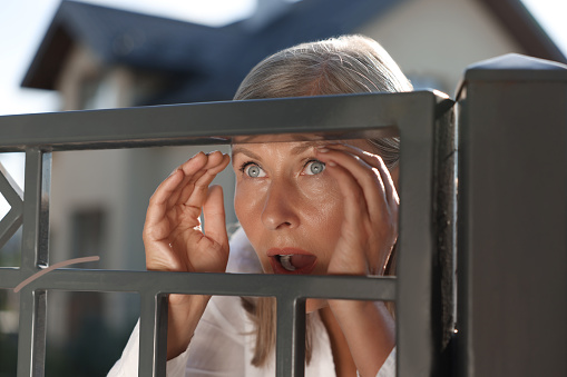 Concept of private life. Curious senior woman spying on neighbours over fence outdoors