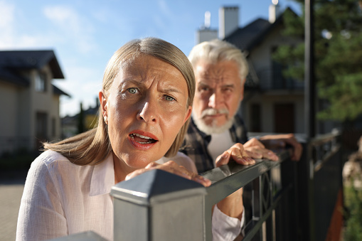 Concept of private life. Curious senior couple spying on neighbours over fence outdoors, selective focus