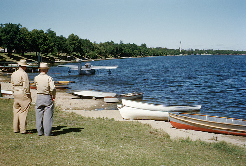 Two men watch a floatplane on a lake at a resort in the late 1940s