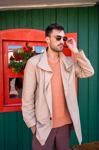 Fashionable man in sunglasses. Stylish and confident male wearing shades, salmon-color sweater and beige trench coat. Portrait of Handsome guy with dark hair standing outside Christmas house.