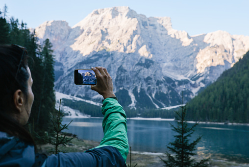 female hiker enjoys the beautiful outdoors in Dolomites, Italy. She is relaxing in the early morning next to Lago di Braies, taking photos with her smartphone.