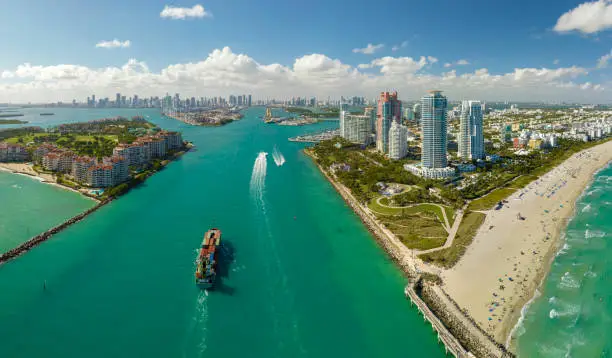Photo of Commercial container ship entering Miami port harbor through main channel near South Beach. Luxurious hotels and residential buildings on waterfront and high skyscraper towers of downtown in distance