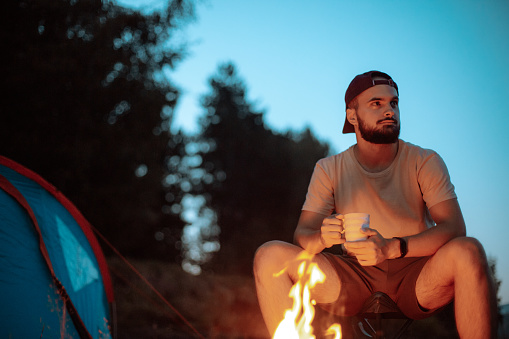 Young hiker man relaxing in nature, he is sitting next to camp tent and fire and drinking coffee