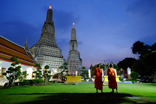 Capture of barefoot Thai monks before dawn in street of Chiang Khan. Group of monks is walking barefoot along street. At right side is a food market stall. A man is kneeling and honoring respect to monks in background waiting for them. Monks are getting food for their day while along street in end of night until sunrise. Monks are wearing traditional orange clothes