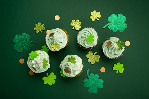 St. Patrick's Day vanilla and chocolate cupcakes with green frosting and  shiny clover decorations on green paper background. Irish holiday dessert concept. Top view, copy space.