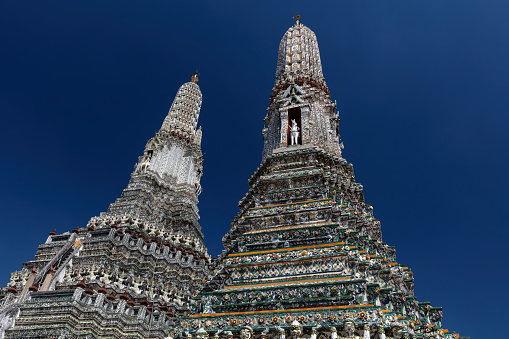 Two prangs at Wat Arun (Temple of the Dawn) in Bangkok, Thailand. Intricate decorations on structures; deep blue sky behind.
