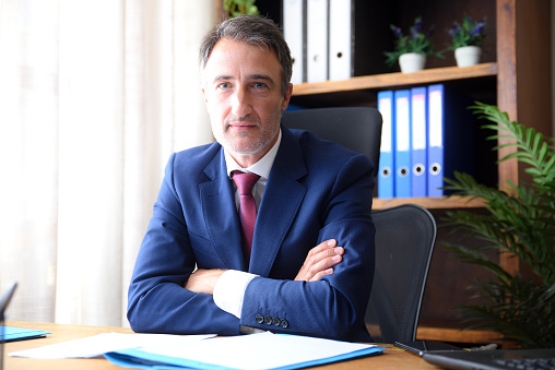 Businessman in his home office wearing blue suit and red tie looking straight ahead with documents on wooden table and bookshelves background.