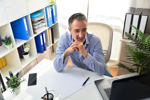 Freelance entrepreneur smiling looking at a laptop in his home office looking proudly at a laptop sitting on a chair in a white office with shelves full of folders. Elevated view.