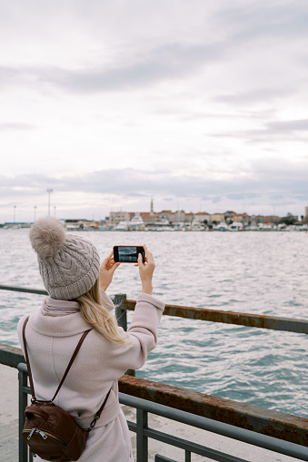 Girl with a backpack stands on the pier at the railing and takes pictures of houses on the seashore with a smartphone. Back view. High quality photo