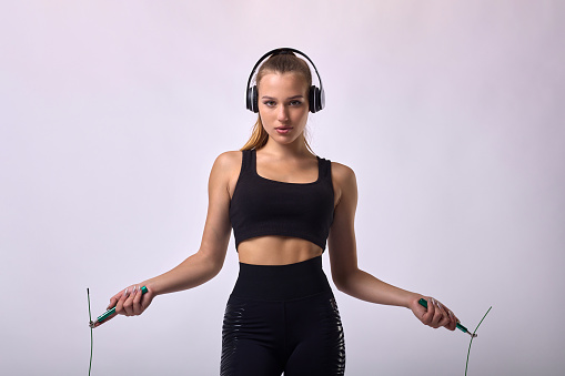 Young beautiful female athlete training with skipping rope, listening to music in headphones during training. Slender sporty beautiful girl in black top jumping on workout