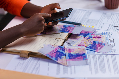 Black person in orange long sleeves shirt working on desk, touching phone screen with notebook and West African CFA franc notes on desk. accounting concept