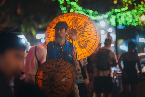 Bangkok, Thailand - January 12, 2024:\nA Thai street vendor sells umbrellas as he walks along busy Khao San Street at night.