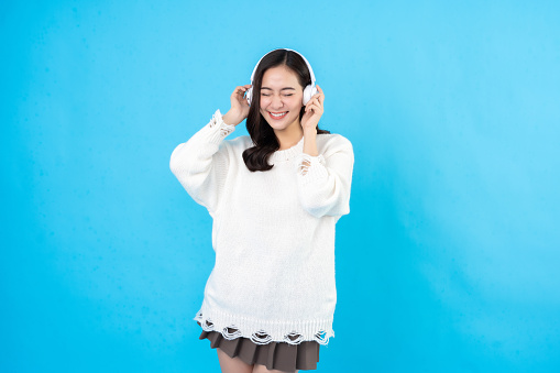 Asian female model Standing as presenter with wireless headphones and listen to music happily with applications in social networks Taking photos in a studio with a blue background Wear a white coat