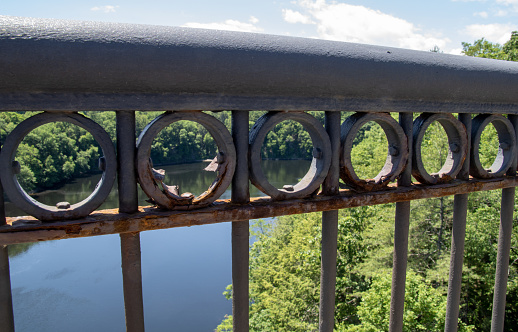 This image shows a series of six rusted metal rings in a guardrail on a bridge in Western Massachusetts. In the background is a calm, serene lake surrounded by lush, green trees.