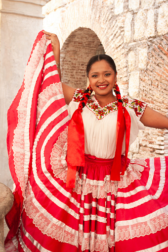 Smiling Mexican woman wearing a traditional dress stands at historical ruins, Oaxaca City, Mexico