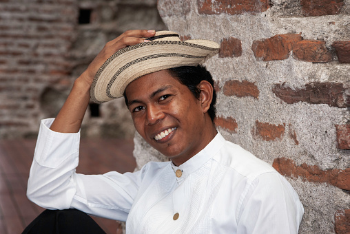 Smiling Panamanian man wearing a traditional white tunic mole and Panama hat sits on ancient stones at Panama la Vieja ruins,  Panama City, Panama, Central America
