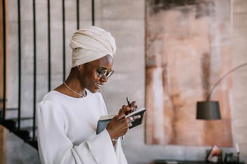 Smiling African woman in white outfit and headwrap writing in notebook indoors