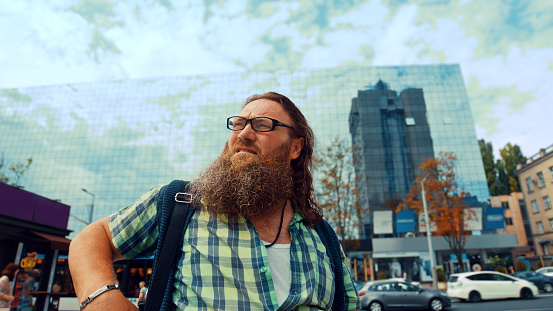 Long-haired man with a beard and eyeglasses worriedly looking away standing in the city square