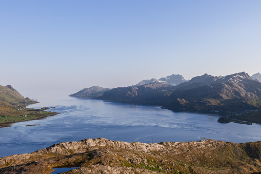 A panoramic view from a high vantage point in the Lofoten Islands, Norway, showcasing the serene Norwegian Sea merging with a dramatic landscape of sharp mountains and a peaceful coastal strip