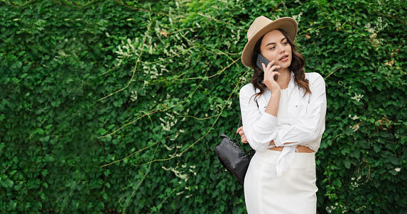 People urban lifestyle concept. Young student woman in straw hat in summer park outdoors rest talk by mobile cell phone look aside. Green nature background