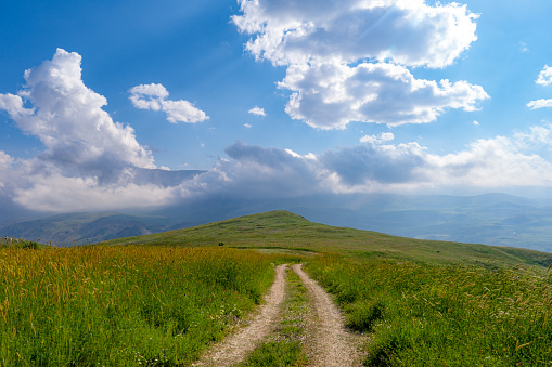 Mountainous region at high altitudes. Meadows, dirt road. Nature and road views. Clear cloudy weather. Sunlight comes through the clouds. Mediterranean region.