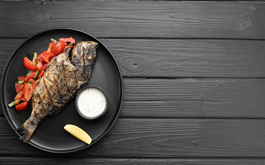 Grilled dorado fish on plate with ingredients over black background, top view. Flat lay. Copy space