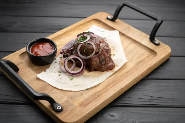 Pieces of pork shish kebab with onions and tomato ketchup served on pita bread with onions on a wooden cutting board. Copy space