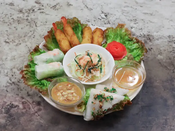 This appetizing image features a Vietnamese Five-Item Cuisine Platter that includes a variety of traditional Vietnamese dishes. Arranged neatly on the platter are fresh and vibrant summer rolls with prawns, golden-fried spring rolls, a tangy noodle salad topped with seafood, delicate steamed rice rolls sprinkled with green onions, and a savory peanut sauce for dipping. The composition is enhanced by a bright, hand-carved carrot rose, all presented on an elegant stone background that brings out the rustic charm of the meal.