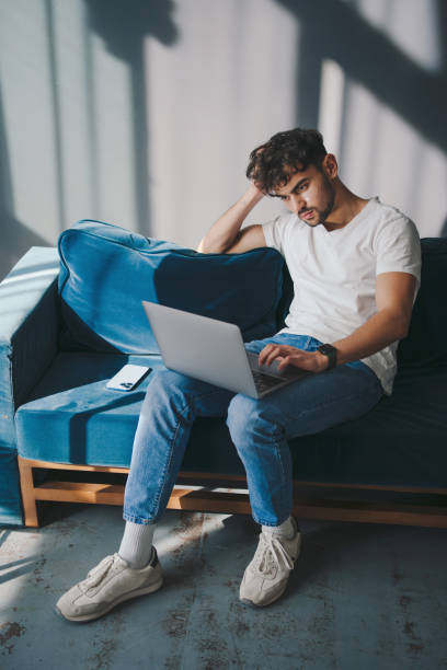 istock Handsome man working on laptop while sitting on couch at home, typing on computer keyboard, enjoying remote work. Cheerful casual male resting on sofa. Enjoying weekend free time. 1947000073