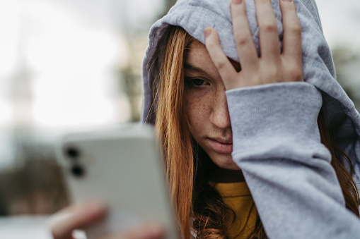 Portrait of teenage girl looking at her smartphone, feeling sad, anxious, alone. Cyberbullying, girl is harassed, threated online.