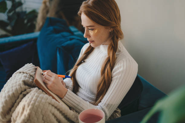 Young redheaded woman sitting on the sofa wraped in a blanket writing notes on notebook with pen. Cozy domestic interior.