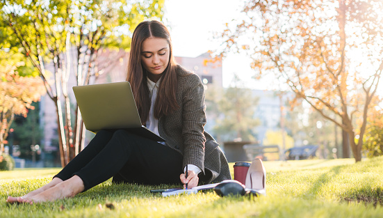 Young brunette woman with laptop sitting on the grass in the park on a sunny using laptop and taking notes. Online training, remote work and communication in social networks