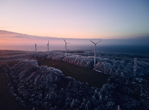 Aerial landscape photography of sunrise over frost-covered nature with wind turbines. Wind turbine towers in soft morning light with icy trees around, harmony of nature and technology. Concept of wind power as clean, renewable energy source.