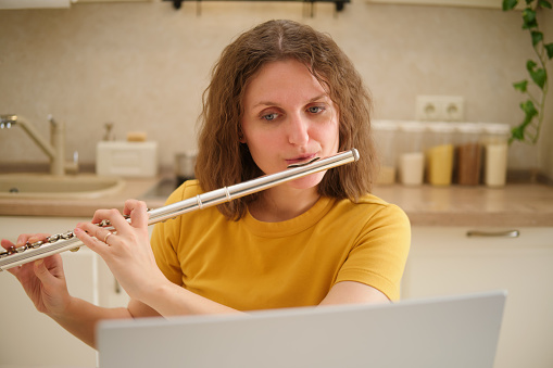 A woman with a laptop playing a flute is sitting at a table in a home kitchen. An adult female businesswoman works from home, a remote office