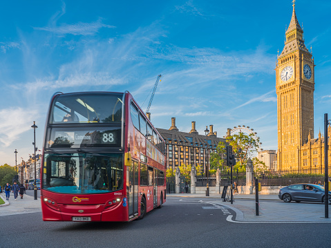 The iconic clock tower of Big Ben and the Houses of Parliament overlooking red double decker bus driving around Parliament Square in the heart of London, UK.
